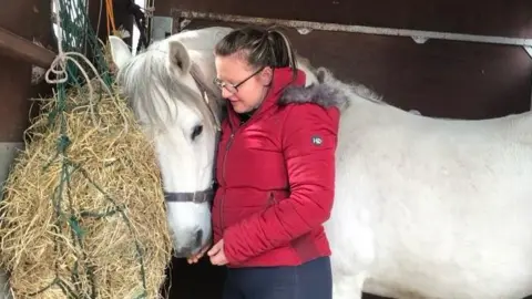 Jennifer Wilson Woman in a red coat hugging a white horse that is standing by a bale of hay in a stable