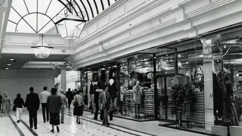 Metrocentre A black and white photo of the shopping centre with people dressed smartly walking down a corridor lined with shops. The central shop has manniquins in the window. The ceiling has an arched window.