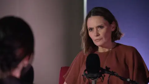 Laura Miller is frowning as she sits in front of a microphone. She is looking intently at a woman she is interviewing - you can only see part of the woman's head from the back