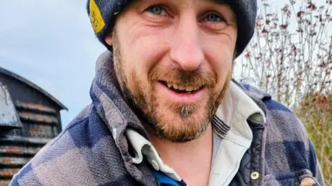 Frankie Colwill A man with facial hair and wearing a hat and checked jacket is pictured close up with a barn and greenery behind him.