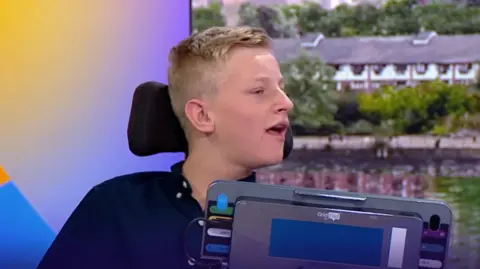 A teenage boy with short blonde hair is in a wheelchair in the BBC Breakfast studio. He is being interviewed and has a tablet in front of him.