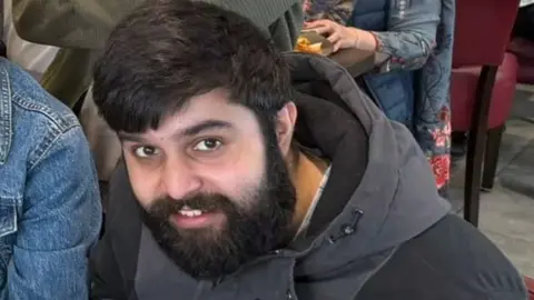 Supplied Adam Ali Hussain, a south Asian man in his 20s with black hair and a medium length beard. He is smiling and looking up towards the frame. He is wearing a black and grey winter coat
