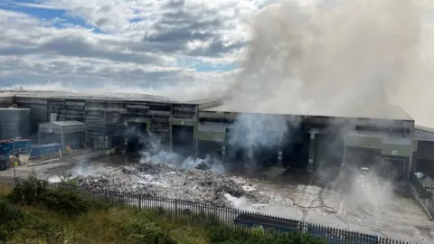 Smoke smoulders across the sites of a warehouse building housing a recycling processing facility with waste mounted up in the yard outside the building and scorch marks across the building.