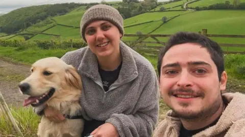 Bethan Mansell A photo of a woman in the middle cuddling a golden retriever. She is smiling and wearing a grey hat and a grey fleece. A man is to the right of her and has dark hair as well as a moustache and beard and has a cream-coloured fleece on. There are fields behind them. The man is taking the selfie.