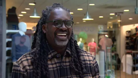 Colin Ayitey - a lan with twisted locs, glasses and a chequered shirt is smiling open-mouthed