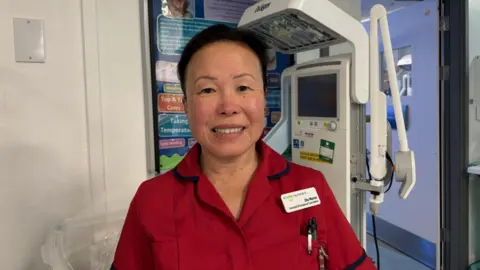 A woman wearing red hospital scrubs smiles for the camera in a clinical setting 