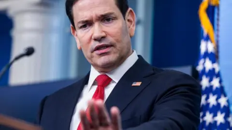 US Secretary of State Marco Rubio speaks at a podium. He is wearing a navy suit and red tie
