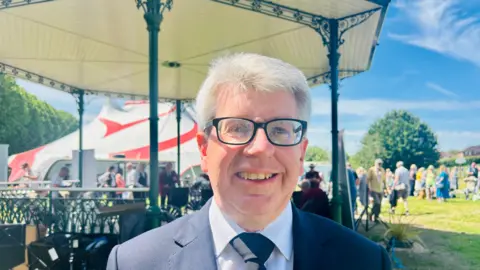 Claudia Sermbezis / BBC A man with white hair and black rimmed glasses looks at the camera and smiles. He is wearing a white shirt, dark tie and dark suit jacket. In the background is a bandstand and a grassy area with people standing on it in the sunshine.