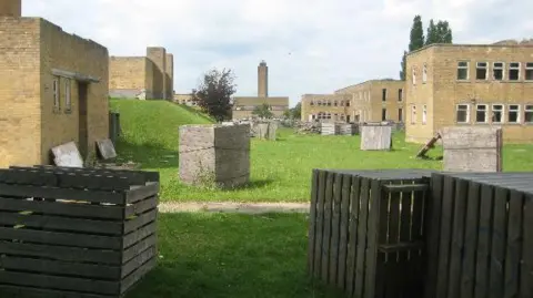 Peter Neal/Geograph Light brown brick buildings - mostly two-storey with small windows. There is a grassed area between the two rows of buildings. Wooden storage boxes are visible at intervals down the grassed area.
