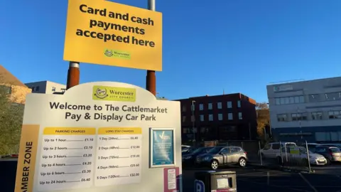 LDRS A sign showing current fees at The Cattlemarket car park in Worcester. There are cars parked in bays in the background, with buildings behind them.