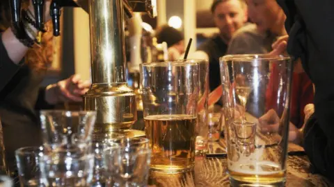Getty Images Pint glasses, shot glasses, and other drinks sit on a busy pub bar counter with people socialising in the background.