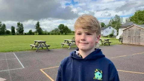 A young boy with blond hair wears a navy blue hoody and stands on the edge of a playing field in a playground. There are picnic benches and a shed behind him.