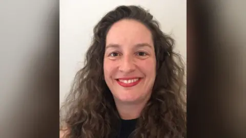 Cambridge University Hospitals Prof Rachel Isba is smiling at the camera while taking a photo of herself against a white background. She has long curly brown hair, brown eyes and is wearing a black top and red lipstick. 