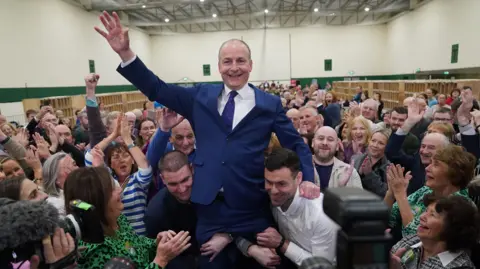 PA Media Fianna Fail leader Micheál Martin is hoisted up by his sons Cillian and Micheál Aodh, after he was deemed elected in the Cork South Central constituency at the election count centre at Nemo Rangers GAA Club in Cork. Crowds of people are around him.