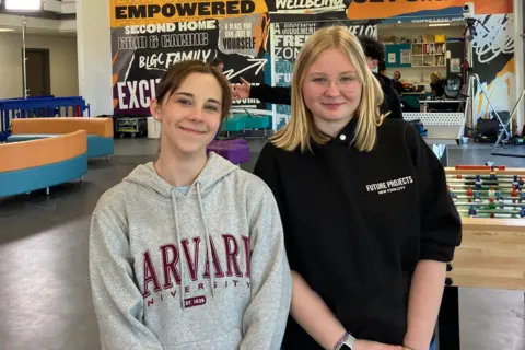 Two girls smiling at the camera as they pose for a picture in a youth club. Maisie on the left has brown hair pulled back in a pony tail and is wearing a grey hoodie with Harvard University branding in large red type. Iga on the right wears a black hoodie bunched up on her arms. She has shoulder-length blond hair and has rimless glasses.
