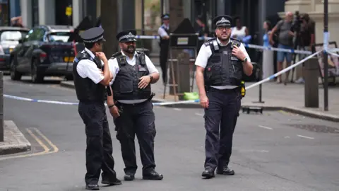 Three police officers are pictured standing on a street in front a police cordon. 
