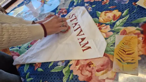 Alix Haigh A woman stamping a design on to a piece of triangular fabric which has the word STATVAM printed on it