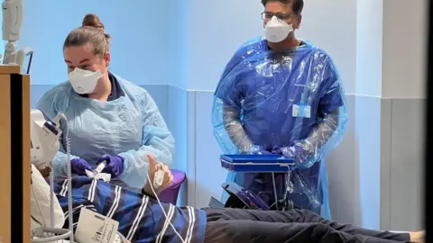 A man lies on the bed with a blue t-shirt with stripes on it - and dark trousers. A woman in PPE looks down on him as he gives a thumbs up. A man is in the background with the same protective gown and both wear white face masks.