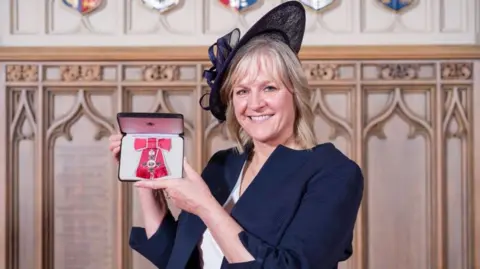 A woman with shoulder-length blonde hair holds up her MBE to the camera, which is a medal on a red bow. She wears a blue jacket and hat.