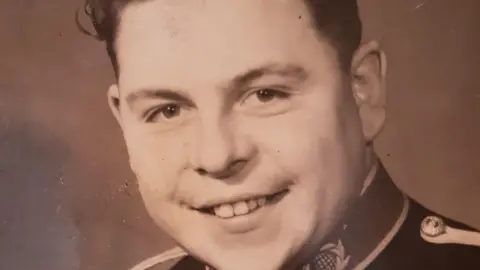 Royal Navy Mr Latham pictured during his time in the Royal Marines. The image is an old photographed that is brown in tone. He is smiling at the camera while wearing his military uniform.