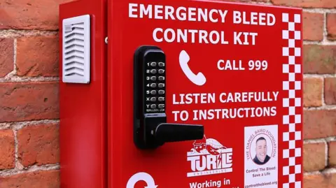 A red metal box on a brick wall. The box has a keypad and handle as well as a phone number and details for those looking to use the bleed control kit.