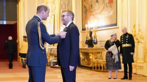 PA Jay Flynn receiving an MBE from Prince William