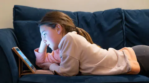A young girl wearing a cream top and grey leggings  is lying on a sofa staring at a brightly lit screen in a dark room 