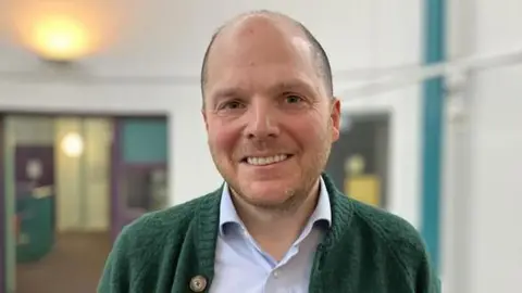 Professor Tom Sperlinger is smiling directly into the camera. He is standing in an empty room with a light behind him. He is a white man, with thinning very short hair, he is unshaven. He is wearing a light blue shirt and dark green cardigan.