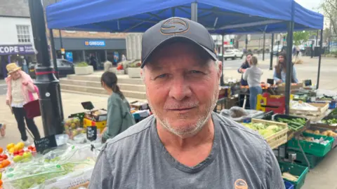 John Devine/BBC Michael Smalley has a stubbly chi and face and a black baseball cap on his head, he is wearing a grey T-shirt. Behind him is a market stall, with produce on display and some customers around it, shops and cars are visible in the background.