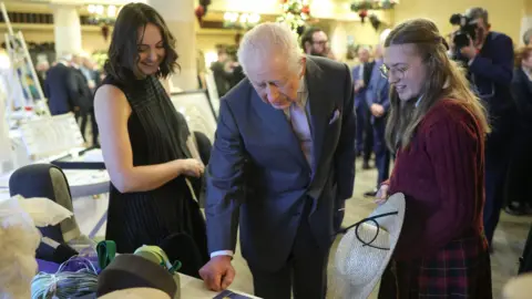 Chris Jackson/PA King Charles III pictured next to two young women. The one to the left of the frame has short dark hair and is wearing black. The woman on the right has long blonde hair, glasses, and is wearing a red dress. King Charles is wearing a dark grey suit jacket and trousers with a white collar shirt and pink tie. All three of them are standing at a table covered with crafts and hats. None of them are looking at the camera - they are looking down at the table.