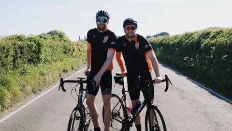 Olly Shone Image shows two brothers smiling at the camera on a country road next to their bicycles. They are wearing matching cycling gear with helmets.