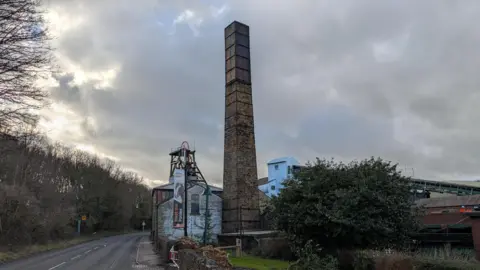 The outside of the National Coal Mining Museum. A tall brickwork shaft stands above a cluster of other buildings around it. A public road runs alongside the site.