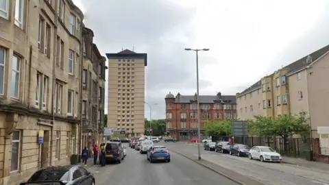 Google A street view of Maxwellton Street street in Paisley, with blocks of tenements and more modern flats either side of a wide road with cars parked on both sides.