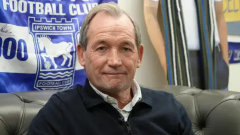 Shaun Whitmore/BBC Burley sits on a grey leather chair with an Ipswich Town flag behind him. He is wearing a blue jacket over a white shirt and is smiling