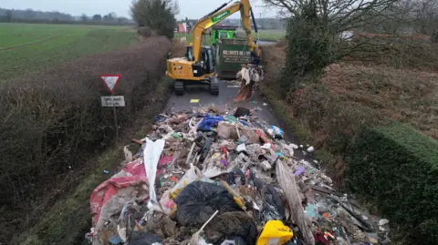 A huge pile of waste in the foreground is being handled by a yellow digger which is scooping the waste into a large green container. The pile of waste visibly blocks the road, and is close to a junction.
