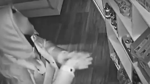 A CCTV image of a young man wearing a hooded jacket and a face covering. He is rubbing his hands together as he looks at a shelf full of sweets.