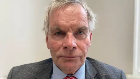 Sharon Edwards/BBC Head and shoulders shot of Martin Hill looking into the camera. He has grey hair and is wearing a blue shirt, red tie and grey suit jacket 