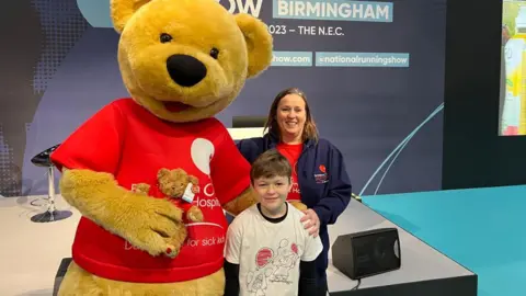 Family A large yellow teddy bear with a red T-shirt next to a small boy with brown hair and a white T-shirt and a woman with brown hair, a red T-shirt and a blue jacket in a room with a blue wall behind them