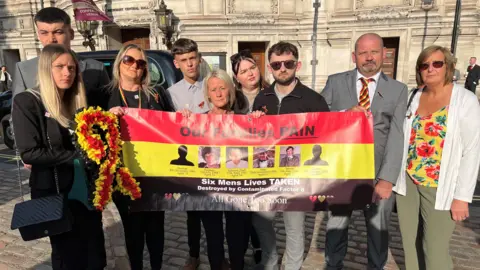 A family is standing in front of a banner which says "our families pain". They are stood in London ahead of the conclusion of the Infected Blood Inquiry. There are nine member of the family ranging in age from their twenties to their fifties. The banner identifies six members of a family that died as a result of infected blood.