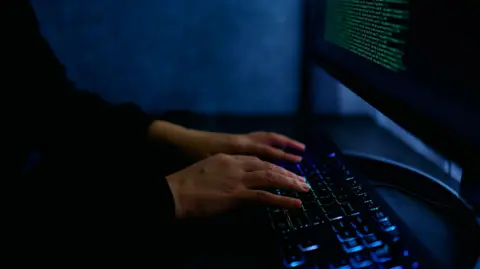 A person types on a lit keyboard in a dark workspace, while a monitor shows green code. 