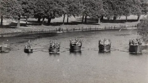Royal Berkshire Archives A black and white photo shows six small boats sailing abreast along a river, with a tree-lined riverbank behind them.