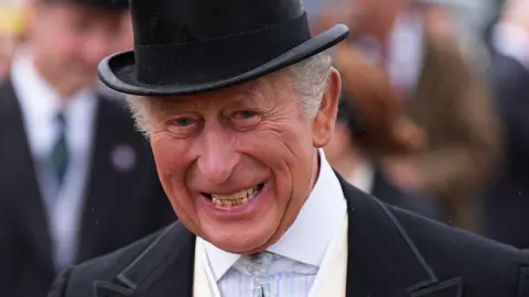 The King, laughing in close-up while wearing a formal morning suit and top hat with a light, patterned shirt and tie. Other people are out of focus in the background.