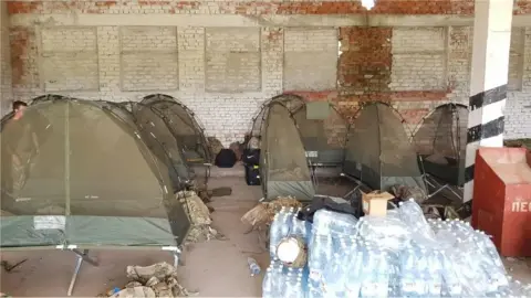 An old abandoned looking building which contains a number of military camp beds which have a tent style netting over each of them. There is a collection of bottled water nearby.