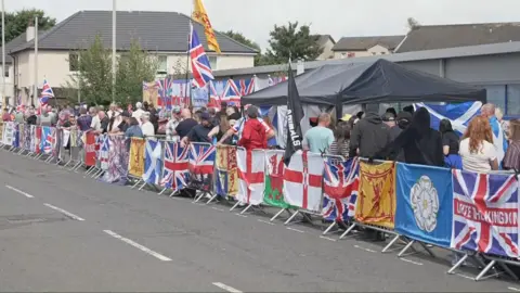 Anti-immigration protesters outside the Cladhan Hotel. They have numerous Union Jack, St George's and Lion Rampant flags draped over the railings they are standing behind and attched to a wall behind them. Others are flying above their heads.