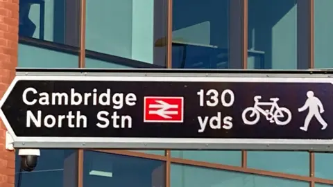 A black sign pointing left says 'Cambridge North Station 130 yards'. There is a red railway logo and an image of a bike and pedestrian. In the background is a red brick building with lots of glass windows. 
