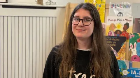 Jen, who has with long brown hair and is wearing glasses and a black jumper, sat down in a playgroup. A shelf full of colourful children's books can be seen behind her. 