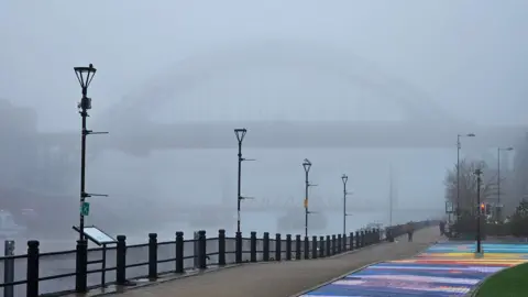 The Tyne Bridge pictured from the Newcastle side in the fog. In the front of the photo is a multi-coloured rainbow path on the quayside. Below the Tyne Bridge you can see the outline of the Swing Bridge and the High Level Bridge. There are multiple lamp posts on the side of the river. 