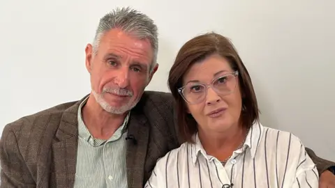 On the left Andrew Bashford has short grey hair with a grey goatee beard. He wears a brown checked jacket over a green striped shirt. On the right Sharon Bashford has shoulder-length dark brown hair. She wears glasses and a white shirt with black stripes.