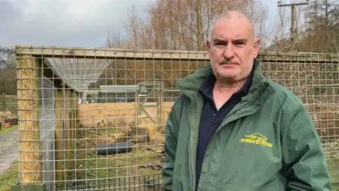 Dave Webb, the founder and CEO of the UK Wild Otter Trust, stood in front of an otter enclosure which is wooden and has metal grated fencing around it. There are trees in the background and the sky is cloudy.