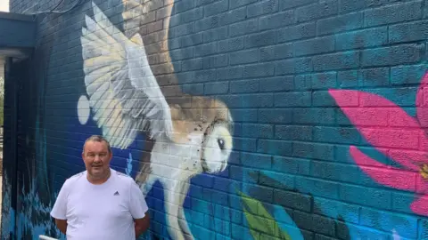 A man with short grey hair is wearing a white t-shirt, he is stood in front of a brightly painted mural on a wall which shows a swooping owl and bright pink flowers